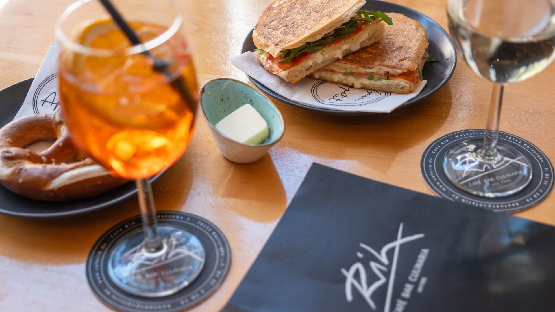 Gedeckter Tisch im Café Rih: Aperol Spritz, Brezel und ein Sandwich auf hellem Holz.